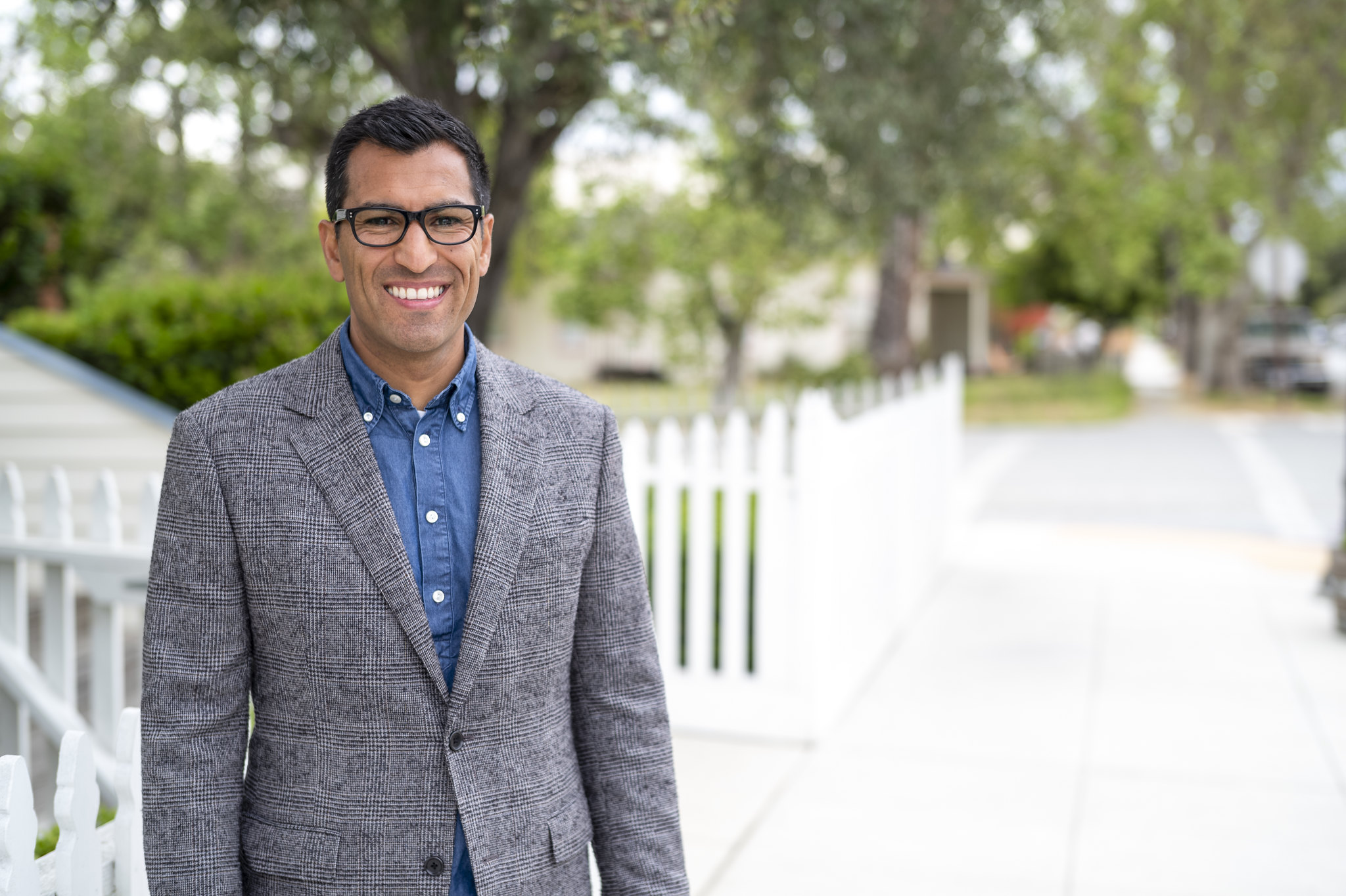 Asm. Robert Rivas pictured on a neighborhood street smiling wearing a blue button up and a dark grey, textured blazer.  